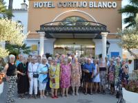 En stor gruppe ældre mennesker står foran et flot hotel i Marbella. Alle smiler og er glade. Solen skinner, det er dejligt vejr, og alle nyder den dejlige dag og hinandens selskab.