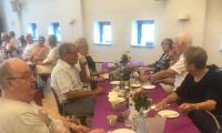 en gruppe ældre mennesker sidder ved et fint dækket bord med blomster, De hygger sig og drikker kaffe
