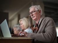 En ældre herre iført en beige cardigan arbejder ved en laptop. I baggrunden sidder to ældre kvinder, der ogås er optaget af at arbejde på hver deres laptop